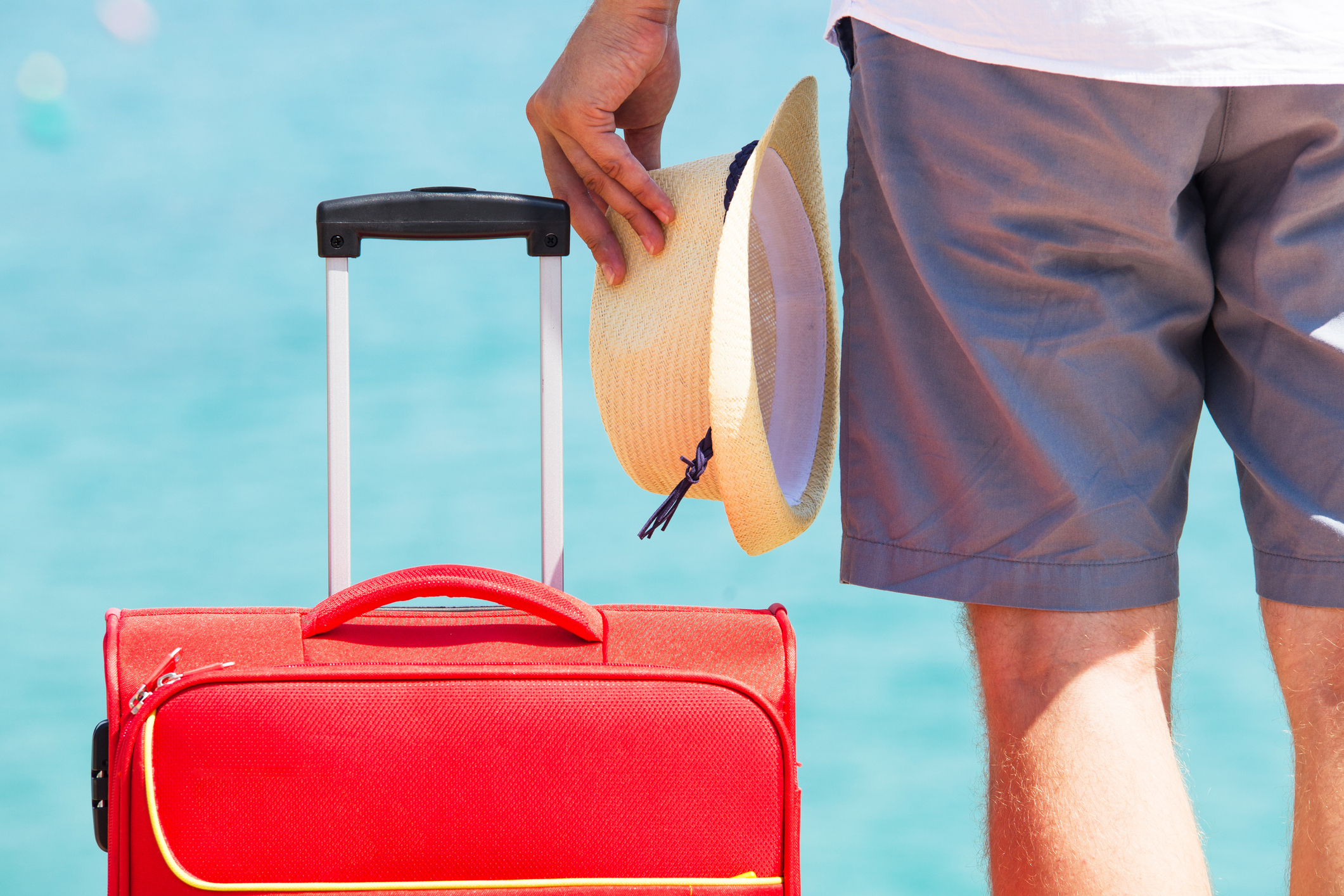 Travel or vacation background. Tropical resort destination scene. Tourist hand holding hat near red suitcase at the sea.